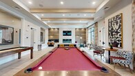 Located at 3 Lexington Ct in Merrimack, NH, this beautifully appointed single-floor amenity space blends contemporary design with a warm, welcoming atmosphere. Light hardwood flooring runs throughout, complementing the exposed wood beam ceiling detail that adds architectural character and depth to the open layout.

At the heart of the space is a generously sized billiards area featuring a striking red-felt pool table set atop a rustic wood frame. A well-equipped wet bar with dark cabinetry and decorative open shelving lines the adjacent wall, ideal for entertaining. Curated artwork adds personality and visual interest throughout.

A stunning double-sided fireplace clad in light natural stone serves as a dramatic focal point, with a mounted flat-screen television above and a sleek dark stone hearth below. Flanking the fireplace is a built-in tufted bench seat and a dedicated business center with workstations, leather chairs, and warm-toned sconce lighting set against rich wood paneling.

The lounge area offers comfortable seating arrangements with patterned accent chairs and side tables. Floor-to-ceiling windows with flowing drapes flood the space with natural light and open to views of the outdoor community, with direct access to an exterior patio. The overall ambiance is sophisticated yet inviting, designed for both productivity and leisure.