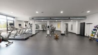 This fitness center is a comprehensive, single-floor facility designed to accommodate a wide range of workout styles and fitness goals. The space is organized into distinct functional zones, all unified by durable dark rubber flooring and crisp white walls that create a clean, professional atmosphere.

The cardio zone features a row of treadmills and elliptical machines, positioned near large windows that allow natural light to fill the space and provide an open, airy feel. An exposed metallic ventilation duct runs overhead, adding an industrial aesthetic element to the otherwise modern interior.

The strength training area is anchored by a multi-station cable machine and a Smith machine, complemented by a full rack of dumbbells and an adjustable weight bench. Floor-to-ceiling mirrors line the walls in this zone, enhancing the sense of space while allowing users to monitor their form during exercise.

A dedicated functional training area offers open floor space equipped with a kettlebell rack holding a variety of weights, exercise mats, foam rollers, and stability balls. A wall-mounted flat-screen television is also present in this area. Amenities include a water station, hand sanitizer dispenser, waste receptacles, and a body weight scale. Recessed lighting throughout ensures consistent, well-distributed illumination across the entire facility.