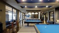 This contemporary amenity lounge is designed to impress, offering a well-appointed social environment spread across a single floor. The space centers around multiple billiards tables set atop bold striped area rugs, creating a defined recreation zone that anchors the room. Overhead, a coffered ceiling with deep navy blue insets and recessed lighting adds architectural depth and visual contrast, while a trio of large, dome-shaped pendant lights illuminates a central lounge and dining area. The lounge seating area features modern blue upholstered chairs and a communal table, ideal for gatherings. Decorative wall art with circular blue motifs adds a cohesive, curated aesthetic to the neutral-toned walls. Polished concrete floors extend throughout, lending an industrial yet refined character to the space. Expansive floor-to-ceiling windows and glass doors on one side flood the interior with natural light while offering views of an adjacent outdoor area, complete with additional seating. A dedicated workspace or meeting nook is visible toward the rear, enclosed with decorative glass panels, providing a functional counterpart to the leisure-focused main area.