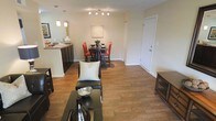 This single-floor residence showcases an open-concept floor plan that integrates the living, dining, and kitchen areas into one cohesive, flowing space. Warm hardwood flooring extends throughout, lending a sense of continuity and understated elegance to the interior.

The living area is anchored by a sleek dark-framed sofa and coffee table, accented by decorative accessories that complement the neutral, cream-toned walls. A track lighting fixture overhead provides focused illumination across the space, enhancing its bright and welcoming ambiance.

Adjacent to the living area, the dining space is defined by a round dark wood table surrounded by bold red upholstered chairs, adding a vibrant contrast to the otherwise neutral palette. Pendant lighting above the nearby kitchen counter reinforces the transition between zones.

The kitchen features dark cabinetry paired with a light stone countertop, offering a striking visual contrast. A pass-through counter connects the kitchen to the dining and living areas, reinforcing the open layout.

Near the entryway, a white front door with clean trim provides a crisp, welcoming entry point. Overall, the space presents a balanced combination of functionality and style, with cohesive design choices throughout.