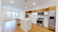 This contemporary single-floor residence showcases a thoughtfully designed open-concept layout that seamlessly blends style and functionality. The kitchen stands as a centerpiece of the home, featuring a two-tone cabinetry design that pairs warm wood-toned upper cabinets with crisp white lower cabinets, complemented by a white subway tile backsplash. A generous central island with a clean quartz countertop provides ample prep and seating space, while stainless steel appliances — including a gas range, built-in microwave, side-by-side refrigerator, and dishwasher — round out the well-equipped culinary space. Light hardwood flooring flows throughout the kitchen and adjoining living and dining areas, lending warmth to the bright, neutral interiors. The living area opens directly to a private balcony through glass-paneled doors, inviting natural light and outdoor views into the space. The bedroom is finished with plush wall-to-wall carpeting and features a dedicated walk-in closet, offering practical storage. The bathroom is cleanly appointed with large-format light stone-look floor tiles, a full soaking tub with a shower, white tiled surround, and polished chrome fixtures, creating a calm and refreshing retreat throughout.