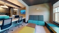 This thoughtfully designed single-floor amenity space combines functionality and modern aesthetics to create an inviting environment for work and relaxation. The space is anchored by a well-appointed business center, where a large marble-surfaced island workstation accommodates multiple desktop computers, complemented by ergonomic mesh office chairs. Elegant globe pendant chandeliers hang from a striking recessed ceiling feature, adding a refined focal point overhead. A wall-mounted flat-screen display and a dedicated printer station further enhance productivity in this area. Adjacent to the workstations, a comfortable lounge zone offers teal and charcoal upholstered seating, colorful accent ottomans, and low wood-finish tables, providing a relaxed setting for informal gatherings. Large windows dressed with patterned valances flood the lounge with natural light and offer views of the surrounding exterior. The kitchenette is outfitted with light wood cabinetry, a sleek quartz countertop, an under-counter beverage refrigerator, a coffee station, and a mosaic tile backsplash that adds texture and visual interest. The entryway makes a strong first impression with bold geometric dark accent lines adorning the walls, large-format tile flooring, and tasteful greenery, setting the tone for the contemporary design language carried throughout the entire space.