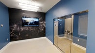 This single-floor space makes a strong visual impression through its thoughtful combination of design elements and functional features. A striking dark accent wall serves as a focal point, adorned with large-scale artistic wallpaper depicting delicate dandelion seed imagery, creating a dramatic contrast against the adjacent blue-painted walls. A wall-mounted flat-screen television is integrated seamlessly into this feature wall, adding a multimedia dimension to the space.

The flooring consists of light-toned tile, contributing to a clean and polished aesthetic throughout. Overhead, fluorescent ceiling fixtures provide bright, even illumination across the room. A set of full-length glass doors with chrome hardware occupies one side of the space, offering both visual transparency and a contemporary architectural detail. The glass enclosure includes what appears to be a mounted control panel, suggesting a functional, tech-integrated design. White baseboards and trim provide clean delineation between the walls and floor, completing the modern, well-appointed character of this space.