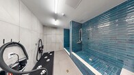Located at 17071 W Dixie Hwy in North Miami Beach, Florida, this purpose-built pet wash station is a well-appointed, single-room facility designed to make pet grooming efficient and comfortable. The space is defined by its striking aesthetic: vibrant teal glazed subway tiles line the walls of the walk-in shower enclosure, creating a bold, eye-catching focal point against the neutral large-format grey ceramic tile flooring and walls that cover the remainder of the room. The open-entry shower stall is fully tiled from floor to ceiling in the signature teal brickwork and is equipped with a flexible chrome spray hose and wall-mounted faucet fixtures, allowing for thorough and easy rinsing. Adjacent to the shower, a cantilevered dark granite grooming bench is set against a matching teal-tiled wall, providing a sturdy elevated surface for post-bath grooming. A professional-grade pet dryer with a flexible hose is mounted nearby, completing the grooming workflow. A paw-print mat on the floor adds a charming, functional touch. The room is brightly lit with overhead fluorescent fixtures and features a clean white ceiling with ventilation. A clearly marked entry door with posted pet wash guidelines ensures ease of use. The overall ambiance is clean, modern, and entirely practical.
