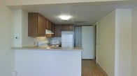 This single-floor space is designed with practicality and functionality in mind. The kitchen serves as the focal point of the layout, featuring dark wood cabinetry that provides generous storage space along the upper walls. A peninsula counter separates the kitchen from the adjoining area, creating a natural division while maintaining an open feel. The kitchen is fully equipped with a white electric range, a built-in microwave, and a full-size refrigerator, making it ready for everyday use. A range hood is mounted above the cooktop, and a sink is conveniently positioned along the counter. The flooring throughout is a warm-toned laminate wood, adding a sense of warmth to the space. The ceiling features a drop-tile panel design with flush-mounted lighting that provides even, ambient illumination throughout. Neutral, light-colored walls keep the space feeling bright and clean. A doorway visible from the kitchen leads further into the unit, suggesting additional rooms beyond. The overall layout is compact yet efficient, offering a well-organized living environment suited for comfortable day-to-day living.