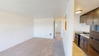 Located at 105 Anoka Ave in Bremerton, Washington, this one-bedroom, one-bathroom unit offers a practical and efficient single-floor layout. The living area is carpeted in a neutral tone and features clean white walls, creating a bright and open atmosphere. A half-wall partition separates the living space from the kitchen, which is equipped with dark wood cabinetry and a gas range, adding a functional contrast to the otherwise light interior. The bedroom is a comfortable size, featuring plush carpet, white walls, and a window fitted with horizontal blinds that allows natural light while maintaining privacy. The window offers a view of the surrounding neighborhood. A hallway connects the main living areas to the bedroom and bathroom, providing a logical and efficient flow throughout the unit. The bathroom includes a full-size tub and shower combination with chrome fixtures and a curved shower rod, complemented by clean, neutral-toned surroundings. Throughout the unit, simple flush-mount lighting fixtures provide consistent illumination. The overall design is straightforward and move-in ready, making it a practical option for those seeking a comfortable space in Bremerton, WA.