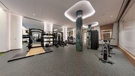 Located at 44 M St NE in Washington, DC, this professionally designed fitness facility spans a generous single floor, offering a comprehensive and upscale exercise environment. The main gym area features durable rubber flooring and is outfitted with an extensive array of equipment, including TRUE-brand cardio machines, a full rack system with TRX suspension trainers, medicine balls, a complete dumbbell rack, adjustable benches, and functional training sleds. A striking dark green tiled column anchors the space visually, complemented by sculptural cloud-shaped recessed ceiling panels with integrated ambient lighting that lend a polished, modern aesthetic. Floor-to-ceiling windows along one wall flood the cardio zone with natural light and offer views to the street, while full-length mirrored walls amplify the sense of space throughout. A separate studio room features warm hardwood flooring, floor-to-ceiling glass partition walls, and a curved architectural niche for equipment storage, making it well-suited for cycling, yoga, or group classes. Additional amenities visible in the space include a sauna room and a connecting hallway. The overall design balances high-end finishes with functional versatility, creating a premium fitness environment.