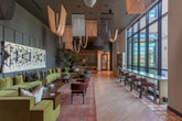 Located at 1200 Rickabaugh Way in Santa Clara, CA, this thoughtfully designed lobby lounge offers a generously proportioned, single-floor open layout that makes a strong architectural statement. Soaring double-height ceilings amplify the sense of volume throughout, while expansive floor-to-ceiling windows along multiple walls flood the interior with abundant natural light and frame views of the surrounding landscape.

The flooring transitions between warm wood-look planks and vibrant geometric patterned tile, adding visual rhythm and defining different functional zones. A central lounge area anchors the space with a sprawling olive-green velvet sectional sofa, layered patterned rugs, organic-shaped wood coffee tables, and a curated textile wall installation that serves as a bold focal point against muted blue-gray paneled walls.

Distinct seating vignettes serve varied needs: a windowed banquette area with built-in bench seating, bistro tables, and woven globe pendant lights provides an ideal setting for casual dining or meetings. A semi-private co-working nook features a shared desk, upholstered stools, and sculpted acoustic wall panels. A dramatic arched booth structure clad in teal upholstery and ornate tilework offers an intimate retreat. Lush tropical plants are positioned throughout, softening the interior and adding a vibrant, resort-inspired ambiance to this dynamic communal space.