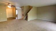 This three-story property offers a straightforward, functional layout with clearly defined living areas across multiple levels. The main floor showcases a spacious carpeted living area finished in neutral beige tones, providing a clean and versatile foundation for furnishing. Olive-green walls run throughout the space, lending a warm, understated character to the interior. A white-painted staircase with open railing leads to the upper floors, positioned centrally to allow easy flow between levels. A floor vent indicates a forced-air HVAC system for climate control. Adjacent to the main living area, a separate room features hard-surface flooring and benefits from natural light through a window, making it well-suited for use as a dining area, office, or additional living space. A doorway with a white door provides access between these areas, maintaining an open yet defined sense of separation. The textured ceiling adds a traditional touch consistent throughout the main level. Overall, the space presents a clean, move-in-ready environment with a neutral palette that accommodates a wide range of interior styles across all three floors.