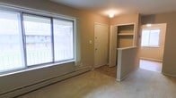 This single-floor unit offers a straightforward, functional layout designed for comfortable everyday living. The main living area is finished with wall-to-wall carpeting, providing a warm underfoot feel, while a large picture window along one wall allows generous amounts of natural light to fill the space and offers a view of surrounding trees and neighboring structures. A baseboard heating unit runs beneath the window, ensuring efficient warmth throughout the room.

Toward the back of the living area, a clearly defined entryway features a solid front door alongside an adjacent built-in shelving niche with open shelves — a practical storage solution ideal for books, décor, or everyday essentials. The transition between the carpeted living space and the entry area is marked by a change to hardwood-style flooring, adding a visual distinction between zones.

A partial half-wall with a small countertop surface separates the living area from an adjacent space, likely a kitchen or dining area, contributing to an open yet organized flow. The walls are finished in neutral warm tones throughout, and a flush ceiling light provides overhead illumination. Overall, the space presents a clean, no-frills environment well-suited for practical, comfortable living.