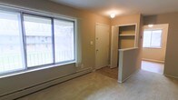 This single-floor unit offers a straightforward, functional layout designed for comfortable everyday living. The main living area is finished with wall-to-wall carpeting, providing a warm underfoot feel, while a large picture window along one wall allows generous amounts of natural light to fill the space and offers a view of surrounding trees and neighboring structures. A baseboard heating unit runs beneath the window, ensuring efficient warmth throughout the room.

Toward the back of the living area, a clearly defined entryway features a solid front door alongside an adjacent built-in shelving niche with open shelves — a practical storage solution ideal for books, décor, or everyday essentials. The transition between the carpeted living space and the entry area is marked by a change to hardwood-style flooring, adding a visual distinction between zones.

A partial half-wall with a small countertop surface separates the living area from an adjacent space, likely a kitchen or dining area, contributing to an open yet organized flow. The walls are finished in neutral warm tones throughout, and a flush ceiling light provides overhead illumination. Overall, the space presents a clean, no-frills environment well-suited for practical, comfortable living.