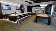 This contemporary amenity lounge is designed with a sophisticated, social atmosphere in mind. The space is anchored by a sleek, elongated wet bar finished in dark wood cabinetry with a crisp white countertop, lined with green-cushioned chrome bar stools that add a lively pop of color. Pendant lighting and recessed fixtures illuminate the bar area, while decorative horizontal ceiling elements add architectural depth and visual interest throughout.

Adjacent to the bar, a regulation-size billiards table with a tan felt surface serves as a central entertainment focal point, inviting friendly competition. Multiple wall-mounted flat-screen televisions are strategically placed throughout the lounge, ensuring clear sightlines from virtually every corner of the room.

A dedicated lounge area toward the rear of the space features comfortable seating arrangements ideal for conversation or casual relaxation. The flooring throughout consists of patterned broadloom carpet in neutral tones, complementing the dark wood accents and light-colored walls. The overall design palette of charcoal, white, and green lends the space a clean, upscale feel. The layout flows openly, making this lounge a well-appointed, multifunctional gathering space suited for a variety of social occasions.