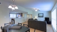 This single-floor residence offers a thoughtfully designed open-concept floor plan that integrates the main living areas into one cohesive, functional space. The kitchen is outfitted with dark cabinetry, a breakfast bar peninsula, and modern stainless steel appliances, with a window above the sink that draws in natural light. The kitchen flows directly into the dining area, which is anchored by a stylish pendant light fixture overhead, creating an inviting space for meals and gatherings. Adjacent to the dining area, the spacious living room features a ceiling fan with a light kit and is furnished with comfortable seating arranged around a central focal point. Soft, neutral wall tones throughout the space lend a calm and airy ambiance, while light hardwood flooring ties the open areas together seamlessly. A landscape-themed framed artwork adds a decorative touch to the living room wall. Interior doors along the back of the space provide access to additional rooms beyond the main living area. The overall layout is practical and well-organized, offering comfortable single-level living with a clean, contemporary aesthetic throughout.