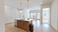 Located at 2124 American Industrial Way in Chamblee, GA, this well-appointed 2-bedroom, 2-bathroom residence offers a thoughtfully designed single-floor layout with modern finishes throughout. The open-concept kitchen and living area serves as the heart of the home, featuring light hardwood-style flooring that flows seamlessly through the main living spaces. The kitchen is outfitted with stainless steel appliances, including a refrigerator, range, and over-the-range microwave. A central island with a quartz countertop, undermount sink, and warm walnut-toned cabinetry is complemented by white upper cabinets with gold hardware and a decorative ogee tile backsplash. Pendant lighting with brass accents adds a refined touch overhead. The living area is bright and airy, with large windows and a door providing access to an outdoor area, filling the space with natural light. Both bedrooms feature plush carpeting and neutral tones, offering a calm retreat. The primary bathroom includes an extended vanity with white cabinetry and ample storage, while the second bathroom features a tub-shower combination with tiled surround. Two closets provide generous storage. The clean, contemporary aesthetic and functional layout make this a comfortable and stylish Chamblee residence.