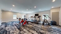 Located at 9250 Shady Lake Dr in Streetsboro, Ohio, this fitness center offers a well-appointed, single-floor workout space designed to support a variety of cardio and exercise routines. The room is finished with neutral, light-toned walls and recessed lighting throughout the smooth ceiling, creating a clean and energizing atmosphere. A bold, abstract-patterned carpet in dark tones with contrasting light streaks covers the floor, adding a dynamic visual element to the space.

The fitness center is equipped with multiple treadmills, an elliptical trainer, a stair climber, and red spin bikes, providing users with a comprehensive selection of cardio equipment. A wall-mounted flat-screen television is positioned for easy viewing during workouts. A large mirror along one wall visually expands the space and allows users to monitor their form.

Natural light filters in through several windows along the perimeter walls, offering views of the surrounding greenery and contributing to an open, airy feel. A dedicated exit door with overhead emergency signage ensures safety and convenient access. The overall layout provides ample open floor space for movement, making this fitness center a functional and inviting amenity for residents of the Shady Lake Dr community.