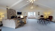 This single-story clubhouse offers a warm and inviting atmosphere designed to accommodate a variety of community activities. The main gathering area is characterized by soaring vaulted ceilings with exposed beams and dual ceiling fans, creating an open, airy feel throughout the space. Plush carpeting in a neutral tone covers the main floor, complementing the cream-colored wainscoting and walls.

A standout feature of the lounge area is the full brick fireplace wall, complete with a mounted flat-screen television, providing a cozy focal point for relaxed gatherings. Comfortable plaid sofas and armchairs are arranged around glass-topped coffee tables, offering ample seating. A built-in window seat beneath a multi-pane bay window allows natural light to fill the room while providing additional casual seating.

High-top tables with bar stools are distributed throughout the space, offering flexible seating arrangements. Deep-seated wingback chairs create quieter conversation nooks along the perimeter. The adjoining kitchen area features a tiled breakfast bar with bar stools, a full-size refrigerator, stove, microwave, and a charming popcorn machine—making it well-suited for hosting events. The welcoming entryway and open layout connect all areas seamlessly.