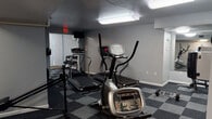 This fitness center is a dedicated, single-floor exercise facility designed to support a well-rounded workout routine. The space is finished with a distinctive checkered gray carpet tile flooring throughout, providing both durability and a clean aesthetic that complements the neutral gray walls and white trim.

The cardio area is well-stocked with multiple treadmills and elliptical machines, arranged to allow comfortable use of each unit. A wall-mounted television is positioned for viewing during workouts, and overhead fluorescent lighting ensures the space is brightly and evenly lit.

A separate strength training zone features a multi-station cable machine, a rowing machine, a weight bench, and a dumbbell rack with a selection of free weights, with a large wall mirror reflecting the equipment and enhancing the sense of space. A sanitation station with hand sanitizer and equipment wipes is conveniently placed near the entrance, encouraging proper hygiene practices.

Natural light filters into the space through vertical-blind-covered sliding glass doors, which open directly to an outdoor area, providing ventilation and a refreshing connection to the exterior. Additional ceiling-mounted fixtures and an exit sign ensure the space remains safe and navigable. The overall atmosphere is practical and functional, offering users a complete environment for both cardiovascular and strength-based training.