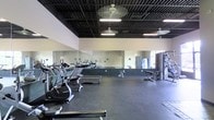 This fitness facility offers a comprehensive and well-organized workout environment designed to accommodate a wide range of exercise needs. The main workout floor is expansive, providing ample space for both cardio training and strength work without feeling crowded.

Along one side of the floor, a row of cardio machines — including treadmills and ellipticals — are neatly arranged for easy access. Strength training equipment and cable machines are positioned toward the rear of the space, offering a variety of resistance training options. The open floor area in between allows for stretching, functional training, or free movement.

A continuous wall of floor-to-ceiling mirrors lines one side of the room, enhancing the sense of space while allowing members to monitor their form during workouts. The dark exposed ceiling adds an industrial character to the space, while ceiling fans and overhead fluorescent lighting ensure the environment remains well-ventilated and evenly lit throughout.

The rubber flooring provides both durability and safety underfoot, making it suitable for heavy equipment and high-traffic use. Natural light filters in through windows along the perimeter, complementing the overhead lighting and contributing to an energizing atmosphere. Overall, the facility is clean, functional, and thoughtfully laid out to support an effective fitness experience.