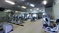 This fitness center is designed to accommodate a comprehensive workout experience within a single-floor layout. The space is generously sized, housing an extensive selection of exercise equipment that includes cable machines, weight stations, recumbent bikes, bench press setups, and various selectorized strength-training machines. The floor is finished with a neutral carpet, providing a comfortable surface throughout the workout area. Mirrored walls line one side of the room, visually expanding the space while allowing users to monitor their form during exercise. The ceiling features multiple ceiling fans and recessed fluorescent lighting panels that ensure the area remains well-lit and ventilated. The overall arrangement of equipment allows for clear pathways and easy navigation between stations. The gray-toned walls contribute to a focused, no-frills athletic atmosphere. Natural light appears to filter in through windows visible in the background, adding brightness to the interior. Overall, the fitness center presents a practical and well-organized environment suited for a full range of strength and cardiovascular training activities.