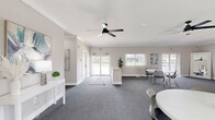 Located at 11 Cinnamon Cir in Randallstown, MD, this single-floor multipurpose space offers a well-designed, open-plan layout ideal for gatherings, meetings, or community events. The interior is finished with neutral warm-gray walls, crisp white crown molding, and clean white baseboards that lend a polished, professional atmosphere throughout.

The main open area is generously sized, carpeted in a durable gray textile, and illuminated by multiple windows fitted with sheer panel shades that diffuse abundant natural light. Ceiling fans with integrated lighting provide both comfort and additional illumination. The space is thoughtfully divided into functional zones: a welcoming lounge area furnished with upholstered sofas and an accent rug, and a flexible meeting or dining zone with round white tables and modern rolling chairs — ideal for collaborative or social use.

A reception or entry area near the front features a white console table and direct access through glass-paneled doors to the exterior. Wall art and potted greenery add warmth and character. Emergency exit signage and accessible features reflect careful attention to safety and compliance. A utility area with printing equipment is also present, supporting office or administrative functions. Overall, the space strikes a balance between inviting comfort and practical flexibility.