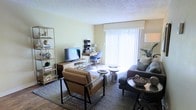 This single-floor space presents a thoughtfully arranged open-concept living and dining area that maximizes both functionality and comfort. The living area is anchored by a plush sofa and a structured leather accent chair, arranged around a pair of round coffee tables atop a patterned area rug, creating an inviting conversational setting. A media console with a flat-screen television serves as a focal point along one wall, complemented by an open shelving unit displaying decorative accessories that add visual interest to the space.

Natural light is a defining feature, pouring in through a large sliding glass door dressed with vertical blinds, which brightens the pale yellow walls and carpeted floors throughout. Adjacent to the living area, a compact dining nook is furnished with a modest table and a modern blue chair, making efficient use of the space near the window. The textured ceiling adds a classic residential character to the room. Overall, the space offers a clean, contemporary ambiance with a warm and livable aesthetic, well-suited for everyday comfort and casual entertaining.