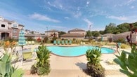 This single-story community amenity facility is defined by its classic brick exterior and welcoming architectural details. The building's entry is framed by a pair of stately wood-framed French doors with multi-pane glass panels and a transom window above, offering a clear sightline to the pool area beyond and flooding the entryway with natural light.

The centerpiece of the outdoor space is a generously sized, freeform swimming pool with striking turquoise water. The pool deck is finished in a warm concrete surround accented with brick-toned edging, and is furnished with an array of lounge chairs and seating positioned for sun exposure and relaxation. Lush tropical plantings, including large-leafed cannas and flowering specimens, are strategically placed around the deck to add color and a resort-like ambiance.

Beyond the pool, a fenced green lawn extends toward a children's playground complete with a colorful play structure and slide, making this an ideal space for families. Tall light poles provide evening illumination throughout the grounds. The surrounding landscape features mature trees and well-maintained shrubbery, creating a pleasant, park-like setting. The brick exterior of the building is accented with traditional lantern-style wall sconces, reinforcing the community's warm, established character.
