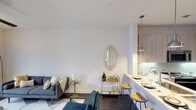 This thoughtfully designed single-floor space in Dallas showcases a contemporary open-concept layout that maximizes both functionality and aesthetic appeal. The living area is anchored by a sleek gray sofa accented with neutral-toned throw pillows, complemented by a glass-topped side table and a decorative gold bar cart beneath an elegant geometric wall mirror — details that add personality and warmth to the space. Clean white walls and smooth ceilings create a bright, airy atmosphere throughout. Transitioning seamlessly from the living area, the kitchen and dining space feature a striking waterfall-edge counter with warm wood bar stools, offering a casual yet sophisticated spot for dining or entertaining. Pendant lighting in a classic industrial style hangs above the counter, casting a warm glow over the space. Light-toned cabinetry lines the kitchen walls, providing ample storage while maintaining the room's fresh, modern character. The flooring shifts subtly between the living and dining zones, delineating each area while preserving the open flow. Throughout the space, a carefully curated palette of whites, warm woods, and gold accents creates a cohesive and inviting ambiance ideal for both everyday living and hosting guests.