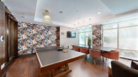 Located at 9 Kimball Ct in Burlington, Massachusetts, this single-floor amenity lounge is a boldly designed, multifunctional space that seamlessly blends recreation, socializing, and relaxation. Rich hardwood flooring runs throughout, anchoring the eclectic yet cohesive interior design.

The most striking architectural feature is a curved, floor-to-ceiling gold chain-link curtain that elegantly divides the lounge into distinct zones while maintaining visual openness. Behind it, a plush curved sectional sofa in charcoal upholstery sits atop a zebra-print rug, flanked by rounded gold coffee tables and illuminated by a sculptural cluster ceiling fixture — creating an intimate, upscale lounge nook.

Bold, large-scale floral wallcoverings in deep navy, blush, and earthy tones adorn multiple walls, infusing energy and personality throughout the space. A full-size ping-pong table serves as the centerpiece of the recreation zone, complemented by a wall-mounted flat-screen television and a stylish wire pendant light overhead.

Adjacent to the game area, a long communal dining table surrounded by plush velvet chairs in warm terracotta tones provides an ideal gathering spot, bathed in natural light from expansive windows that offer views of lush greenery outside. Pendant lighting with gold accents cascades above the table, adding warmth and sophistication. An elevator vestibule with dark wood-framed glass doors provides convenient access to the rest of the building.