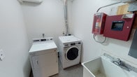 This single-floor laundry facility is purpose-built to meet practical, everyday needs with a clean and functional layout. The room houses a set of commercial-grade laundry appliances, including a front-loading washing machine and a top-loading dryer, both designed for reliable, high-capacity use. A dedicated utility sink is positioned nearby, offering added convenience for hand-washing, pre-treating garments, or general cleaning tasks. Ventilation is efficiently managed through a visible exhaust duct routed along the wall and ceiling, ensuring proper airflow for the dryer unit. Safety is a clear priority in this space, as evidenced by the prominently wall-mounted fire extinguisher housed in a red safety enclosure, making it immediately accessible in the event of an emergency. The room features neutral white walls and a low-profile, easy-to-maintain floor surface that is practical for a high-use laundry environment. The overall design is straightforward and utilitarian, prioritizing functionality and ease of use. This laundry room serves as a well-equipped, self-contained space that provides all essential amenities for efficient laundry management.