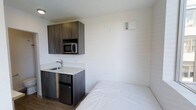 This compact studio unit offers a thoughtfully designed single-floor layout that maximizes every square foot of available space. The open-concept living area seamlessly integrates the sleeping and kitchen zones, creating a functional and cohesive environment. The kitchenette is outfitted with dark wood-toned cabinetry, a stainless steel microwave, a compact refrigerator, and a single-basin sink with a modern faucet, all set against clean white countertops that contribute to a bright, contemporary feel. Wood-look flooring runs throughout the main living area, adding warmth and visual continuity. The walls are finished in crisp white, enhancing the sense of openness within the space. A wall-mounted HVAC unit ensures year-round climate control. The sleeping area accommodates a full-sized bed, positioned to make efficient use of the available floor space. The unit also includes a full bathroom, rounding out the self-contained layout. Clean lines, neutral tones, and purposeful design choices make this studio a practical and comfortable living option suited for individuals seeking an efficient, low-maintenance space.