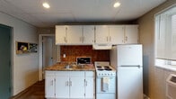 This single-floor unit offers a practical and comfortable layout across several distinct areas. The kitchen is well-equipped, featuring white cabinetry — both upper and lower — with sleek bar pulls, a granite-look countertop with an integrated stainless steel sink, a white electric coil range, a full-size refrigerator, and a bold terracotta tile backsplash that adds a warm, distinctive character. A drop tile ceiling with recessed lighting keeps the space feeling bright and functional. The open living and dining area transitions naturally from the kitchen, offering room for a sofa, coffee table, and a compact dining set. Textured walls and a drop tile ceiling continue throughout, with a window allowing natural light into the space. The bedroom is a quiet retreat with painted cinder block and textured walls, a drop tile ceiling with recessed lighting, and warm wood-patterned flooring. The bathroom is clean and efficiently designed, with white subway tile surrounding the tub and shower, a shower curtain enclosure, tile flooring, a toilet, and a vanity. Overall, the unit offers a cohesive, functional living environment with a casual, lived-in ambiance suited for everyday comfort.