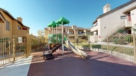 This outdoor community recreational space is thoughtfully designed to serve residents of the surrounding multi-story residential buildings. The centerpiece of the area is a vibrant, multi-feature playground structure topped with distinctive green canopy-style roofs. The play structure includes staircases, slides, a climbing wall, tunnel elements, and interactive panels, all set atop a cushioned rubber safety surface in a deep red hue that provides both safety and visual appeal.

The playground is fully enclosed by metal fencing, ensuring a secure environment for children. Adjacent to the play area, wooden picnic benches offer a comfortable spot for residents to gather and supervise. A concrete pathway connects the various sections of the space, providing accessible circulation throughout.

Beyond the playground, a gated pool area is visible, complete with lounge chairs, a patio table with an umbrella, and a trash receptacle, creating an inviting setting for relaxation. The surrounding residential buildings, finished in a warm stucco exterior with balconies, staircases, and landscaped greenery, frame the space and contribute to its community-oriented atmosphere. The entire area benefits from abundant natural light and clear, open skies, enhancing the welcoming and active character of this shared outdoor amenity.