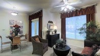 This single-floor space exudes a warm and refined character, thoughtfully designed to balance comfort and style. The living area serves as the focal point of the space, drawing the eye with rich, dark burgundy drapes and valances that frame multiple windows, allowing generous amounts of natural light to filter through. A classic ceiling fan with integrated lighting anchors the room from above, providing both functionality and a traditional aesthetic touch. French doors open to the outside, enhancing the sense of openness and connectivity to the outdoors while adding an architectural charm to the interior. The room is furnished with wicker and upholstered seating, complemented by dark wood accent furniture, including a substantial console piece that adds depth and contrast against the neutral-toned walls. Decorative lamps, candles, and carefully arranged accessories contribute to a layered, curated ambiance. Soft, neutral carpeting underfoot ties the space together, creating a cohesive and inviting atmosphere throughout. Overall, this space offers a classic, comfortable environment with thoughtful details and a timeless decorative sensibility that makes it feel both lived-in and welcoming.