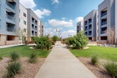 Located at 4910 W Glenn Dr in Glendale, Arizona, this thoughtfully designed outdoor courtyard space serves as a central hub for the surrounding multi-story residential community. The grounds are framed by modern, multi-story buildings with clean contemporary lines, neutral stucco exteriors, and private balconies that overlook the landscaped common areas below.

A wide, concrete pedestrian pathway forms the spine of the courtyard, connecting various outdoor amenity zones and inviting leisurely strolls through the grounds. The landscape design strikes a balance between lush green lawn areas and desert-adapted plantings, featuring ornamental grasses, native shrubs, and mature trees that provide shade and visual interest throughout.

Multiple seating areas are distributed across the courtyard, including shaded covered lounge spaces furnished with wood-and-cushion outdoor furniture, as well as open-air dining table groupings ideal for gathering. A striking sculptural installation featuring colorful bird figures serves as an artistic focal point within the central plaza area.

Additional amenities include a fenced dog park with dedicated turf, raised concrete planter beds suited for community gardening, a picnic table area, and decorative pergola structures. Low-profile pathway lighting ensures the space remains functional and welcoming into the evening hours. The overall ambiance is open, community-oriented, and well-maintained, reflecting a vibrant outdoor living environment in the Arizona sunshine.