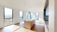 This contemporary single-floor residence is defined by its clean, minimalist architecture and an abundance of natural light. The open-plan living and kitchen area serves as the heart of the home, anchored by a sleek central island with light wood cabinetry and a quartz countertop. The kitchen is equipped with an induction cooktop, wall oven, and crisp white upper cabinets, all set against a seamless white backsplash. Tall, dark-framed floor-to-ceiling windows wrap the living space, framing sweeping panoramic views of the surrounding cityscape and rolling hills beyond.

Exposed concrete structural columns add an industrial counterpoint to the otherwise bright, white-walled interiors, while wide-plank light hardwood flooring runs continuously throughout. The bedrooms are generously proportioned, each benefiting from large windows that capture urban views — including a striking mural visible from one room — as well as accent concrete wall panels that echo the building's architectural character.

The bathrooms are finished to a high standard: one features a glass-enclosed walk-in shower with large-format stone-look tile and mosaic floor detailing, while the other offers a tiled soaking tub and shower combination. A dedicated in-unit laundry closet with a stacked washer and dryer adds everyday convenience. The hallway features an exposed concrete accent wall, reinforcing the cohesive modern aesthetic throughout.