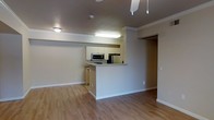 This single-floor space showcases an open-concept design that seamlessly connects the living area and kitchen. The kitchen is defined by a raised peninsula counter that provides both a functional workspace and a natural divider between the cooking and living zones. White cabinetry lines the upper walls of the kitchen, complemented by stainless steel appliances including a microwave and refrigerator, offering a clean and modern aesthetic. Recessed and flush-mount lighting fixtures illuminate the kitchen area effectively, while a ceiling fan with pendant-style lights serves the main living space. Crown molding runs along the ceiling perimeter, adding a refined architectural detail throughout. Light-toned hardwood-style flooring extends across the entire main area, contributing to a bright and cohesive feel. The walls are finished in a neutral gray-beige tone, providing a versatile backdrop. A doorway visible from the main living area leads to an additional room, suggesting a functional layout with private spaces separate from the common living areas. White baseboards frame the flooring throughout, completing the clean, well-maintained interior.