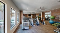 The fitness center at 6047 Hulen Street in Fort Worth, Texas, is a well-equipped exercise facility housed in a single-room space designed to support a comprehensive workout routine. The room features a warm, industrial aesthetic defined by striking exposed brick walls that contrast with a clean white ceiling fitted with recessed lighting and a ceiling fan for optimal air circulation and comfort during workouts.

The exercise room is stocked with a solid selection of cardio machines, including Matrix-brand treadmills, ellipticals, and stationary bikes, arranged to maximize the use of the available floor space. Full-length mirrors line portions of the walls, visually expanding the room while allowing users to monitor their form during exercise. A wall-mounted flat-screen television provides entertainment during workouts.

For strength training, a rack of dumbbells spanning a range of weights is positioned along the brick wall, accompanied by a weight bench. Additional fitness accessories, including medicine balls, resistance bands, and stability balls, are neatly organized on a storage rack, offering variety for functional training routines.

Large windows along one wall allow natural light to fill the space and provide views of the outdoor surroundings. Durable patterned carpet covers the floor throughout. A glass-paneled door provides direct outdoor access, contributing to the room's open and welcoming atmosphere.