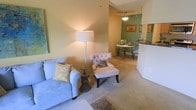 This single-floor residence showcases an open-concept layout that seamlessly connects the living and dining areas, creating a cohesive and functional flow throughout the space. The living area is finished with plush wall-to-wall carpeting in a neutral tone, providing a warm and comfortable atmosphere. Soft, warm-hued walls complement the overall inviting ambiance, while a tall floor lamp adds a layer of ambient lighting to the space.

The living area comfortably accommodates a light blue sofa accented with decorative pillows, paired with a tufted accent chair — both centered around a decorative side table, creating an intimate seating arrangement. A patterned area rug anchors the space subtly.

Transitioning toward the back of the unit, the dining area is distinguished by a sage green accent wall that adds a pop of color and visual interest. A round dining table with chairs sits beneath a pendant light fixture, creating a defined dining space. Adjacent to the dining area, a partial view of a counter — likely a kitchen bar or pass-through — is visible, suggesting convenient access between the kitchen and dining zones. Framed artwork on the walls adds a finishing decorative touch throughout.