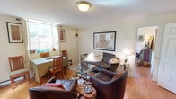 This single-floor space integrates a living area and dining nook into a cohesive, functional layout with warm hardwood flooring throughout. The living area is anchored by rich brown leather sofas and armchairs arranged around a glass-topped coffee table with an ornate metal base, creating an inviting gathering space. A framed artwork piece serves as a focal point on the main wall, complemented by a classic table lamp that adds ambient lighting alongside the central flush-mount ceiling fixture. A decorative floor lamp with an intricate design adds character to the corner near the seating area. The dining nook, positioned near a window, features a green-draped table paired with sturdy wooden mission-style chairs, bathed in natural light from the street-facing window. White walls keep the space feeling bright and open, while thoughtful décor elements — including small plants on the windowsill and decorative objects on surfaces — lend a lived-in, personalized character. A hallway visible to one side suggests additional rooms beyond, completing this compact yet well-appointed single-level layout.