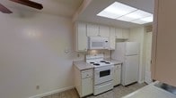 This single-floor space offers a clean, practical layout designed for comfortable everyday living. The main living area is notably open and airy, featuring large sliding glass doors dressed with vertical blinds that allow natural light to filter into the room. The floors throughout are finished with wood-look planking that provides durability and a neutral aesthetic, complementing the warm beige walls and textured popcorn ceilings.

The kitchen is efficiently designed, making smart use of available space with white cabinetry extending to the upper portion of the walls for ample storage. A built-in over-the-range microwave, a coil-burner electric range, and a full-size refrigerator make up the appliance suite, all finished in white for a cohesive look. Light-toned laminate countertops provide functional prep space. An overhead panel light fixture brightens the kitchen workspace effectively.

The overall ambiance of the space is understated and move-in ready, with a neutral color palette of whites and soft beiges that creates a versatile backdrop. Clean baseboards, standard electrical outlets, and straightforward finishes throughout reflect a space that is practical, low-maintenance, and well-suited for a variety of living arrangements.