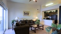 This single-floor residence offers a thoughtfully designed layout that balances comfort and functionality. The living area serves as the heart of the home, boasting impressively high ceilings that lend an airy, spacious feel to the room. A ceiling fan overhead provides both style and practicality, while warm, neutral-toned walls create an inviting atmosphere throughout. Soft, plush carpeting underfoot adds a cozy element to the space, complementing the richly toned furnishings. A decorative framed landscape painting adorns the wall, adding a touch of character to the living room. The space flows naturally into the kitchen, which is visible through an open pass-through counter, featuring dark cabinetry and modern appliances, including a built-in microwave and refrigerator. This open-concept connection between the living and kitchen areas enhances the sense of space and encourages easy interaction. Ambient lighting from tasteful table lamps, combined with overhead lighting, creates a warm and welcoming glow throughout. The overall ambiance of this residence is one of relaxed comfort, with a neutral palette and classic design elements that make it both livable and visually appealing.