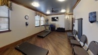 This fitness center offers a comprehensive and welcoming environment for a variety of workout routines. The space is finished with rich hardwood-style flooring, warm neutral walls, and elegant dark wood crown molding that lends a refined character uncommon in typical gym settings. Decorative valance window treatments frame multiple windows throughout, allowing natural light to filter in and brighten the workout area.

The main exercise area is equipped with a Matrix treadmill, a SCI FIT recumbent cycle, and a stability ball station, providing options for both cardio and low-impact training. Exercise mats are available for floor work and stretching, while resistance bands mounted to a dedicated wall rack support strength and flexibility training. A ceiling fan and flush-mount lighting keep the space comfortable and well-lit throughout the day.

A wall-mounted flat-screen television adds convenience for entertainment during workouts. Full-length mirrors along one wall enhance the sense of space and allow users to monitor their form. Seating chairs are arranged along the perimeter, providing a designated rest area. The layout is open and functional, accommodating both individual exercise sessions and small group activities with ease. Overall, the fitness center balances practicality with a warm, inviting aesthetic.