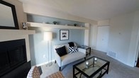 This single-floor space centers around a thoughtfully designed living area that combines comfort with contemporary style. The room is finished with soft, blue-grey walls that create a calm and inviting atmosphere, complemented by white trim and a textured ceiling that adds subtle visual interest overhead.

A standout architectural feature is the built-in horizontal display shelf that spans one wall, positioned at an elevated level. This shelf provides both a decorative and functional element, currently styled with small potted plants and decorative objects, drawing the eye upward and adding dimension to the space.

The living area is furnished with a tufted light grey loveseat accented by black and patterned throw pillows, paired with a sleek glass-top coffee table with a dark metal frame. A floor lamp provides warm ambient lighting, enhancing the overall coziness of the room. Carpeted flooring runs throughout, adding warmth underfoot.

A sliding or paneled closet door is visible along one wall, suggesting practical built-in storage. The overall ambiance of the space is modern, clean, and well-organized, with a cohesive neutral and muted tone palette throughout.