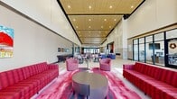 This expansive single-floor community space offers an impressive and welcoming environment defined by its dramatic soaring ceilings and warm wood-paneled ceiling accents with recessed lighting throughout. The open-plan layout is anchored by a grand entrance with floor-to-ceiling curtain-wall glazing, flooding the interior with abundant natural light and providing sweeping views of the surrounding landscape.

The main lounge area features bold, vibrant red and pink upholstered sofas, armchairs, and sectionals arranged across large-format stone tile flooring, complemented by striking abstract area rugs in rich red and black tones. Colorful artwork adorns the walls, adding an artistic character to the space. A dedicated game area includes a full-sized billiards table alongside casual seating, while a secondary lounge zone offers a mounted flat-screen television and additional comfortable seating arrangements. A bar-height countertop area with upholstered stools and a wall-mounted display provides a focused casual workspace or social gathering spot with direct outdoor views. Throughout the space, intimate conversation clusters with armchairs and accent tables create versatile zones suited for both social gatherings and quiet relaxation, making this a well-appointed and dynamic communal environment.