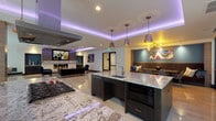 This single-floor space is a modern, open-concept amenity area designed with an upscale aesthetic and a seamless flow between functional zones. The expansive layout is anchored by polished large-format tile flooring that reflects the ambient lighting, contributing to the space's refined atmosphere.

The kitchen area is a standout feature, showcasing two substantial dark-cabinetry islands topped with striking veined granite countertops. A professional-grade gas cooktop, integrated microwave, and a stainless steel range hood overhead underscore the space's culinary capabilities. Pendant lights in muted purple and copper tones hang above the islands, complementing a continuous LED cove lighting system installed along the tray ceiling perimeter, which casts a dramatic purple glow throughout.

Adjacent to the kitchen, the lounge area is anchored by a sleek linear gas fireplace set into a textured stone surround, topped by a large wall-mounted display screen. Curated seating—including a rich brown leather sofa and deep navy accent chairs—surrounds a sculptural round coffee table, creating an inviting conversational setting.

A dedicated dining nook with upholstered booth seating and decorative artwork provides an intimate dining option. Floor-to-ceiling windows along one wall invite natural light and outdoor views, enhancing the open, airy character of the space.
