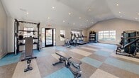 Located at 1970 Peachford Rd in Dunwoody, GA, this single-floor fitness center offers a generously sized, thoughtfully designed space for comprehensive workouts. The main gym area is bright and open, featuring vaulted ceilings with recessed lighting and a ceiling fan that contribute to an airy, energizing atmosphere. Large windows along multiple walls invite abundant natural light and provide views of the surrounding outdoors, enhancing the overall workout environment. The flooring throughout the gym consists of a modern, multi-toned carpet tile pattern in shades of blue, gray, and beige, adding visual interest while providing comfort underfoot. The facility is well-stocked with a wide range of equipment, including multiple treadmills with integrated displays, elliptical machines, a stationary bike, cable and weight machines, an extensive dumbbell rack, adjustable benches, and stability balls. Full-length mirrors line one wall, amplifying the sense of space and allowing users to monitor their form. Two bathrooms serve the facility, each finished with neutral hexagonal tile flooring and clean white walls. The bathrooms are ADA-accessible, featuring open vanities with modern fixtures, framed mirrors, and glass-partitioned shower and stall areas. The overall design is clean, modern, and functional, making this fitness center a practical and inviting space for daily exercise.