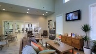 This single-floor amenity space seamlessly blends relaxation and fitness into one well-designed environment. The lounge area commands immediate attention with its soaring double-height ceilings, which lend a sense of openness and grandeur to the space. Recessed lighting illuminates the room evenly, complementing the natural light that filters in through large windows fitted with blinds. A wall-mounted flat-screen TV adds a modern, functional touch to the lounge setting.

The seating area is thoughtfully arranged with a plush sofa accented by colorful throw pillows, paired with two leather accent chairs and a wooden coffee table — creating an inviting atmosphere ideal for socializing or unwinding. A rustic wooden credenza along one wall provides additional surface space and storage, while decorative accents add warmth to the neutral gray-toned walls.

Adjacent to the lounge, the fitness center is visible through a wide open doorway, offering a range of strength and cardio equipment including cable machines and ellipticals. Ample natural light flows into the fitness area as well, creating a bright and energizing workout environment. Together, these two spaces form a cohesive and functional amenity area built around community comfort and wellness.