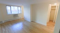 This single-floor space offers a clean, move-in-ready interior with a simple and functional design. The main living area is finished with light-toned hardwood flooring that runs throughout, lending warmth and continuity to the space. Smooth, cream-colored walls provide a neutral backdrop that pairs well with a variety of furnishing styles. Natural light enters through a window fitted with horizontal blinds, brightening the room while allowing for privacy control. A wall-unit air conditioner is installed beneath the window, supplemented by baseboard heating that runs along the perimeter of the room — ensuring year-round comfort. The open layout includes a partial wall or alcove area that adds a subtle sense of room division, offering flexibility for defining distinct living zones without the use of full walls. Clean white baseboards frame the flooring throughout, adding a polished finishing touch. The overall ambiance is understated and versatile, well-suited for those looking to customize a tidy, well-maintained space. Electrical outlets are conveniently positioned along the walls to accommodate various furniture arrangements and functional needs.
