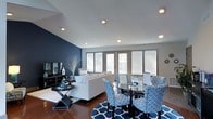 This single-floor space showcases a thoughtfully designed open-concept layout that combines a living area and dining space into one cohesive environment. The most striking architectural feature is the deep navy blue accent wall, which anchors the living area and adds a bold contrast to the surrounding light gray walls and crisp white ceiling. Recessed lighting is strategically placed throughout the ceiling, providing even, ambient illumination across the entire space.

The living area is furnished with a plush, neutral-toned sofa accented with blue and white decorative pillows, complemented by a sleek, low-profile media console along the accent wall. Rich hardwood flooring runs throughout, adding warmth to the cool color palette.

The dining area features a round glass-top table surrounded by patterned blue and white upholstered chairs, positioned atop a coordinating geometric area rug that ties both zones together visually. Large windows with white horizontal blinds line the far wall, allowing natural light to filter in while maintaining privacy. A set of glass doors further enhances the brightness and offers an outdoor connection, making the space feel open and airy.