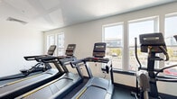 Located at 4121 Concord Creek Place in Richmond, VA, this single-floor fitness center offers a thoughtfully organized workout space designed to support a range of exercise routines. The room features durable rubber flooring throughout, providing a safe and functional surface for all types of physical activity.

The cardio zone is prominently positioned along the windowed wall, which features multiple large windows that flood the space with natural light and offer views of the surrounding community. This area is equipped with a row of treadmills, upright stationary bikes, and an elliptical machine, all from Matrix — a well-regarded commercial fitness brand.

On the opposite side of the room, a strength training area includes a multi-functional cable and pulley rack, a weight bench, free weights, and colorful exercise mats for floor-based workouts. A large wall-mounted mirror enhances the sense of space and allows users to monitor their form during strength exercises. Recessed lighting supplements the natural light, ensuring the entire room is well-illuminated at any time of day.

A bold teal accent wall adds a pop of color to the otherwise clean, neutral palette of white walls and gray flooring, giving the space a modern and energetic aesthetic. A sanitizing station and waste bin are conveniently provided, reflecting attention to cleanliness and user comfort.
